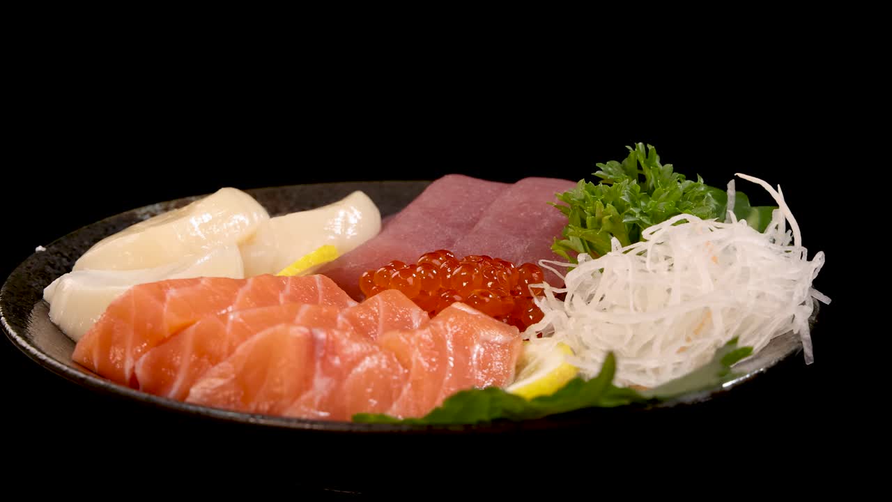 A plate of assorted sashimi and sushi, including salmon, tuna, scallop, and roe, rotates smoothly under bright studio lighting against a black backdrop