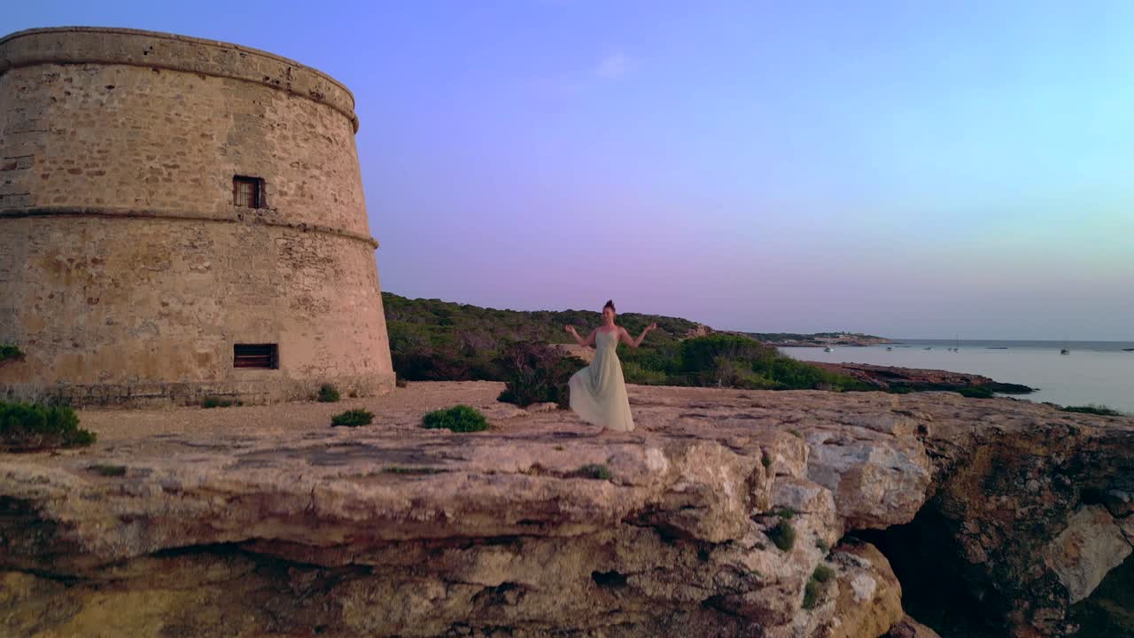 Barefoot hippie girl practicing yoga at sunset near Torre des Carregador, Ibiza, Spain. speed ramp hyper motion time lapse. Great aerial view flight descending drone
