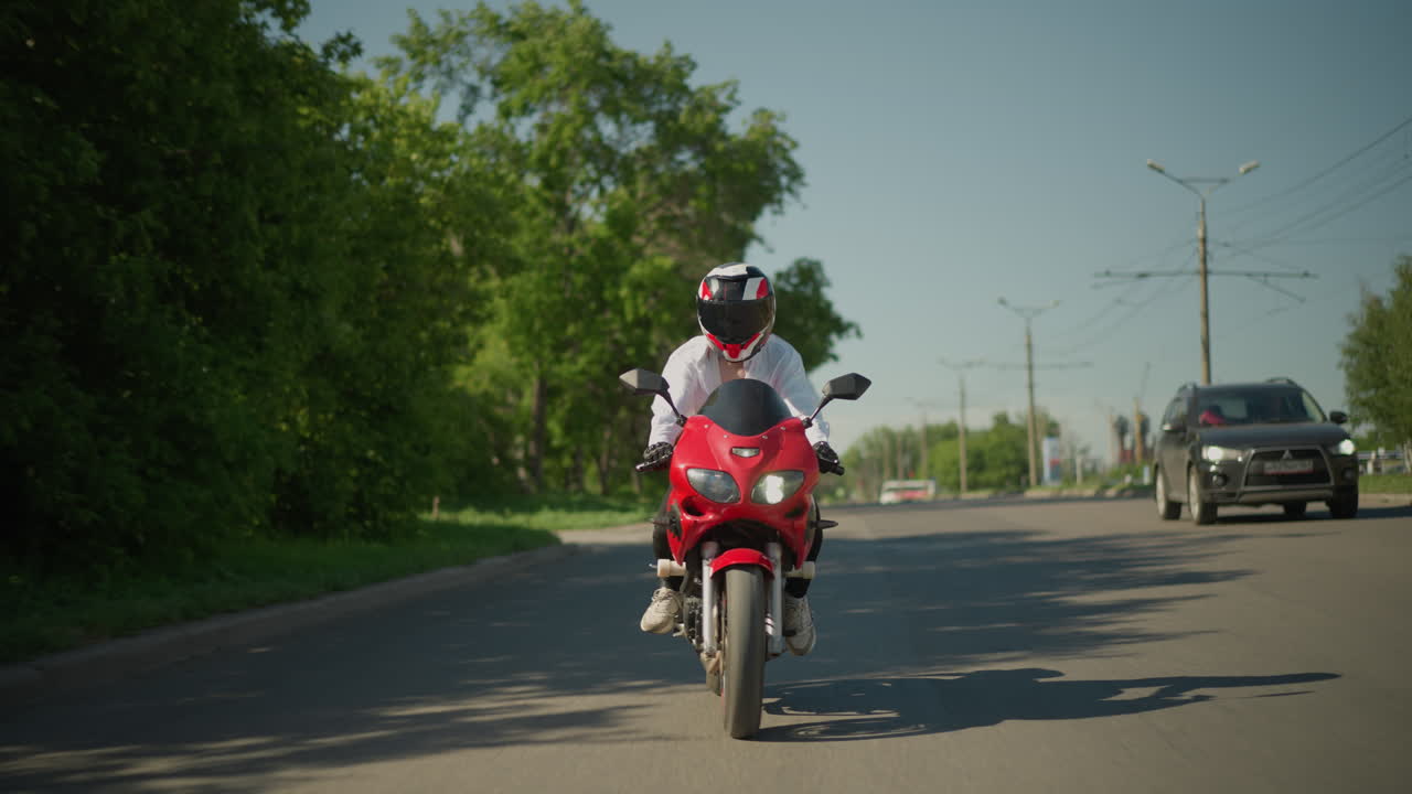 una motociclista femenina en una bicicleta eléctrica roja cruza un carril peatonal mientras un coche se mueve a su lado en un área urbana, el fondo incluye árboles verdes, postes eléctricos
