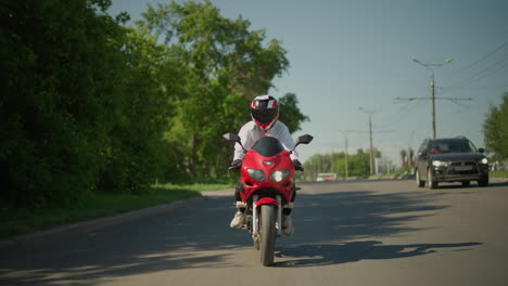 a female motorcyclist on a red power bike crosses a pedestrian lane while a car moves beside her in an urban area, the background includes green trees, electric poles