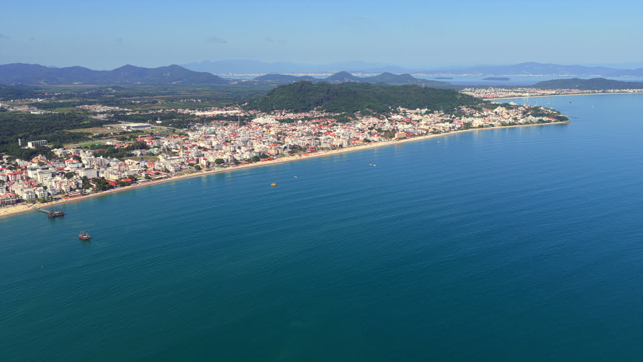 Ascesa aerea elevata con allontanamento sopra l'oceano blu, che guarda verso fitti edifici e verdi colline di Canasvieiras, Florianópolis.
