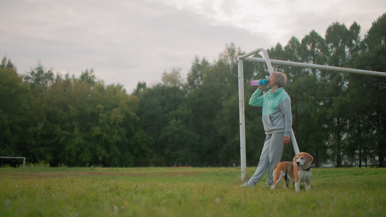 Graceful woman in mint and gray activewear relaxing after morning exercise, drinking from colorful water bottle while standing next to her playful beagle puppy on grassy sports field under cloudy sky