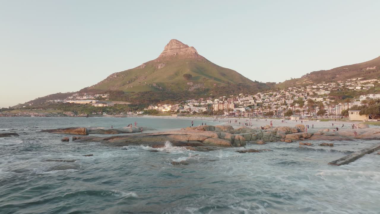 Drone flies backwards over the beach of Camps Bay in Cape Town, South Africa - in the background the Lion's Head Mountain rises illuminated by the sunset - people on the beach