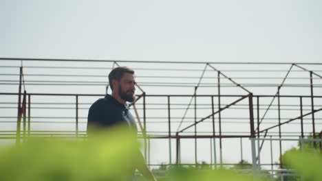 close up green leaves in foreground with side view of agronomist walking and pausing to inspect farmland near metal frame structure during sunny field evaluation