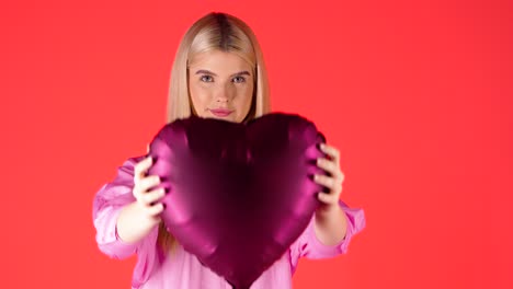 Mujer-Rubia-Bonita-Sonriendo-Sosteniendo-Un-Globo-En-Forma-De-Corazón-Púrpura-Contra-Fondo-Rojo,-Foto-De-Estudio