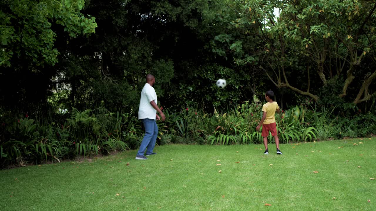 African American father retrieving soccer ball from shrubs, tossing pass to teenage son, copy space