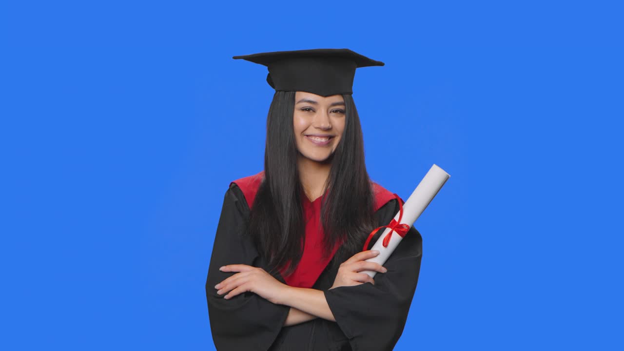 retrato de una estudiante en traje de graduación de gorra y bata sonriendo a la cámara y sosteniendo su diploma. mujer joven posando en el estudio con fondo de pantalla azul. primer plano. cámara lenta lista 59.94fps