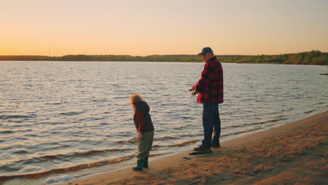 old-fisher-is-catching-faith-in-river-by-fishing-rod-his-little-grandson-is-playing-on-sandy-shore