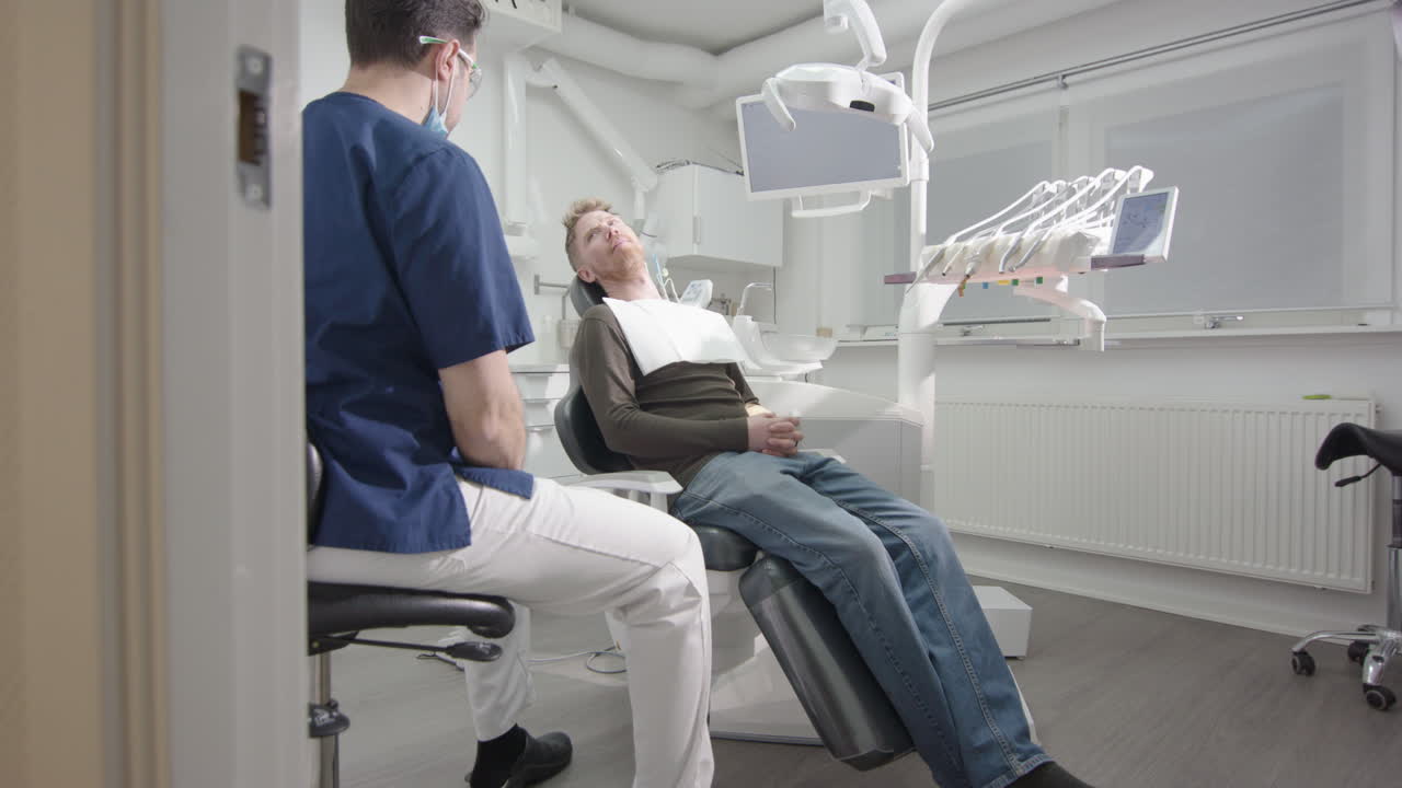 Male patient in reclined chair listens to prognosis from dentist after oral exam