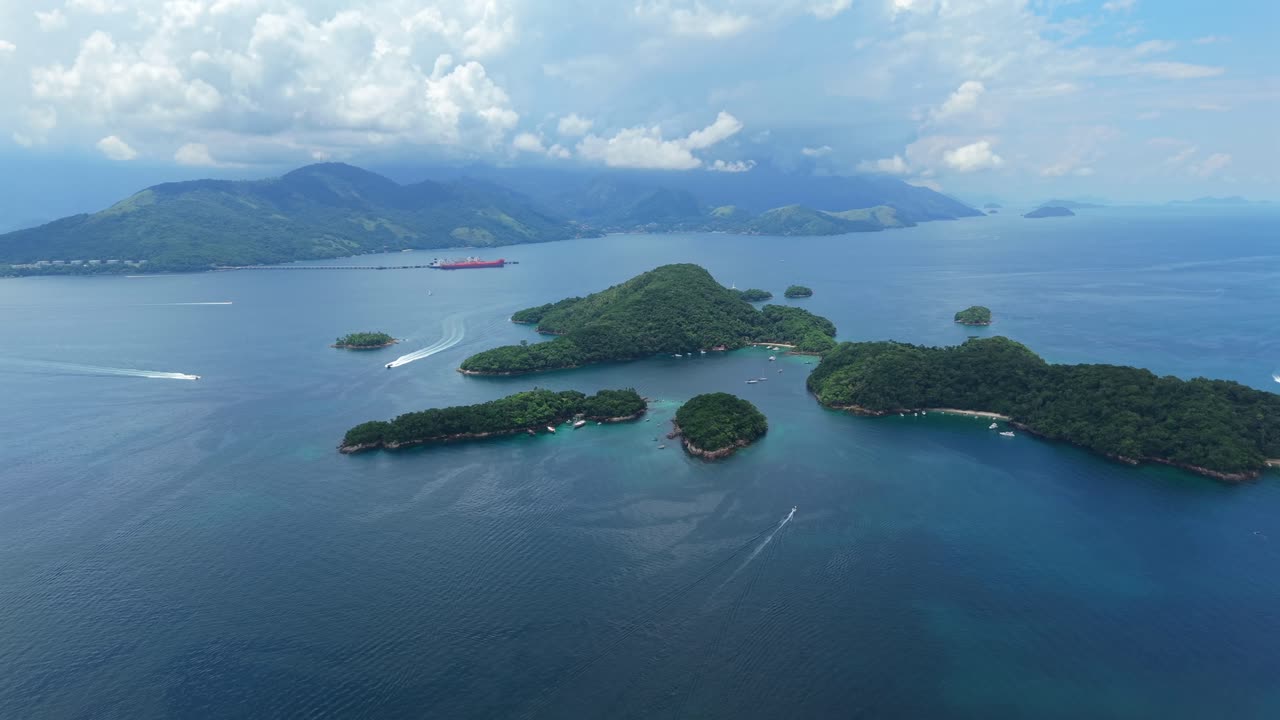 Gugusan pulau bervegetasi dan samudra di Brasil pada hari tropis yang berawan, dengan beberapa perahu melintasi air biru. Pemandangan udara sudut lebar yang cepat dalam 4K