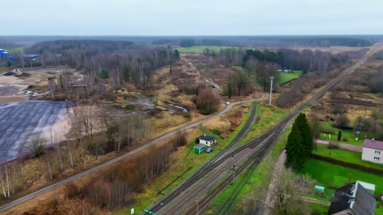 Vista aerea di binari ferroviari, una stazione rurale e campi autunnali circostanti che si estendono verso una foresta distante e strutture industriali in una giornata nuvolosa