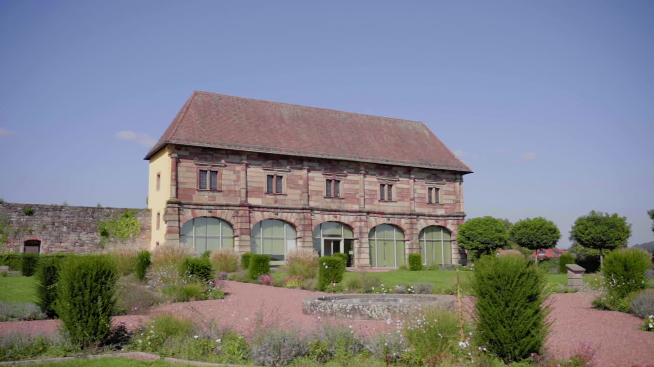 This is a slow-motion shot of the historic Orangery in Blieskastel, featuring traditional architecture and a lush summer landscape. It is a captivating scene of greenery and a blue sky.