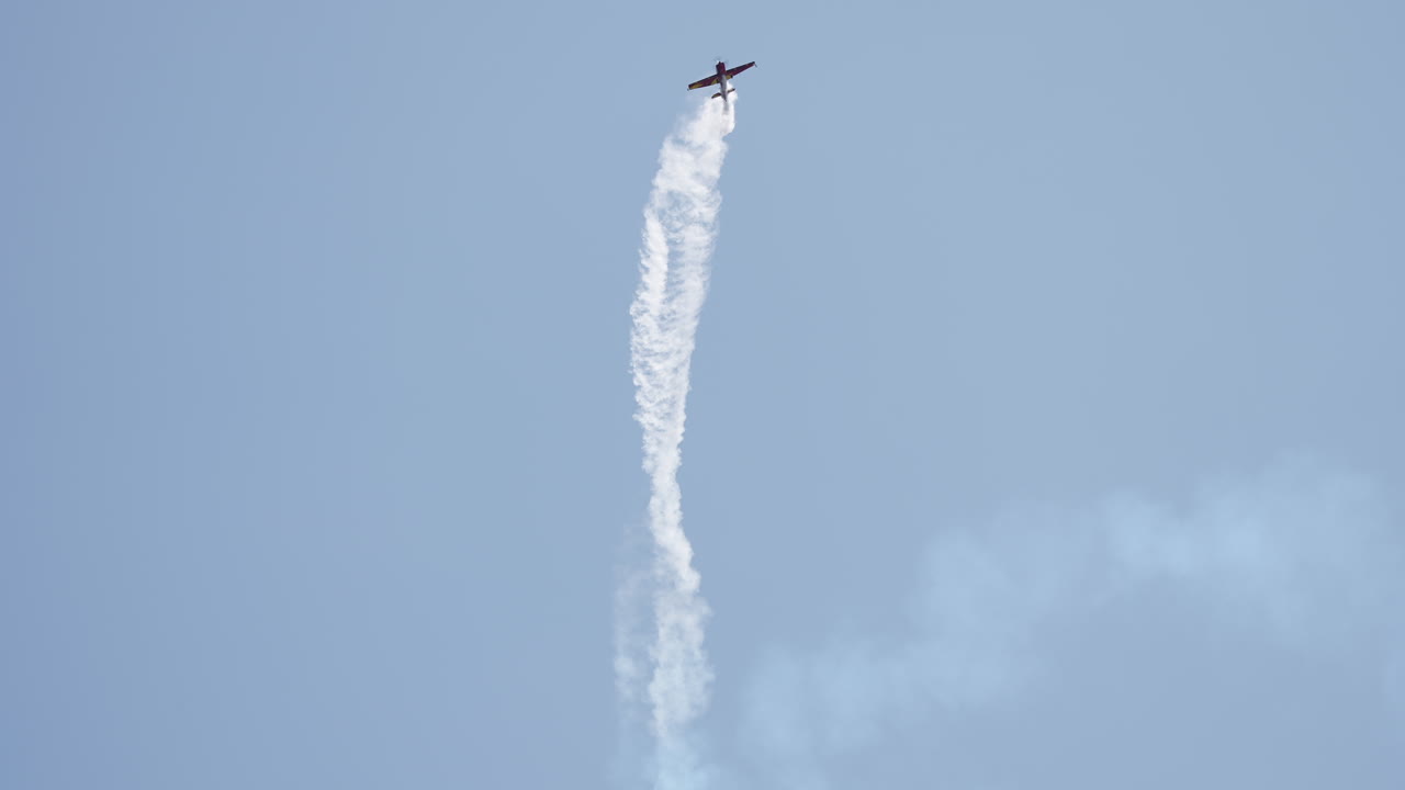 Brightly painted aerobatic airplane flies low over hills while turning and leaving smoke trail behind