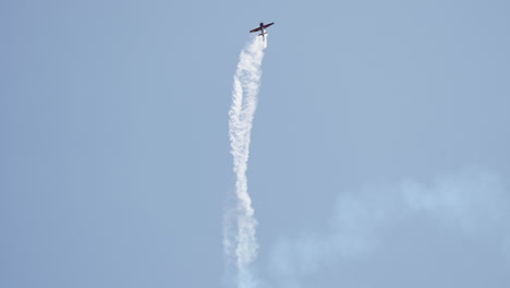 brightly painted aerobatic airplane flies low over hills while turning and leaving smoke trail behind