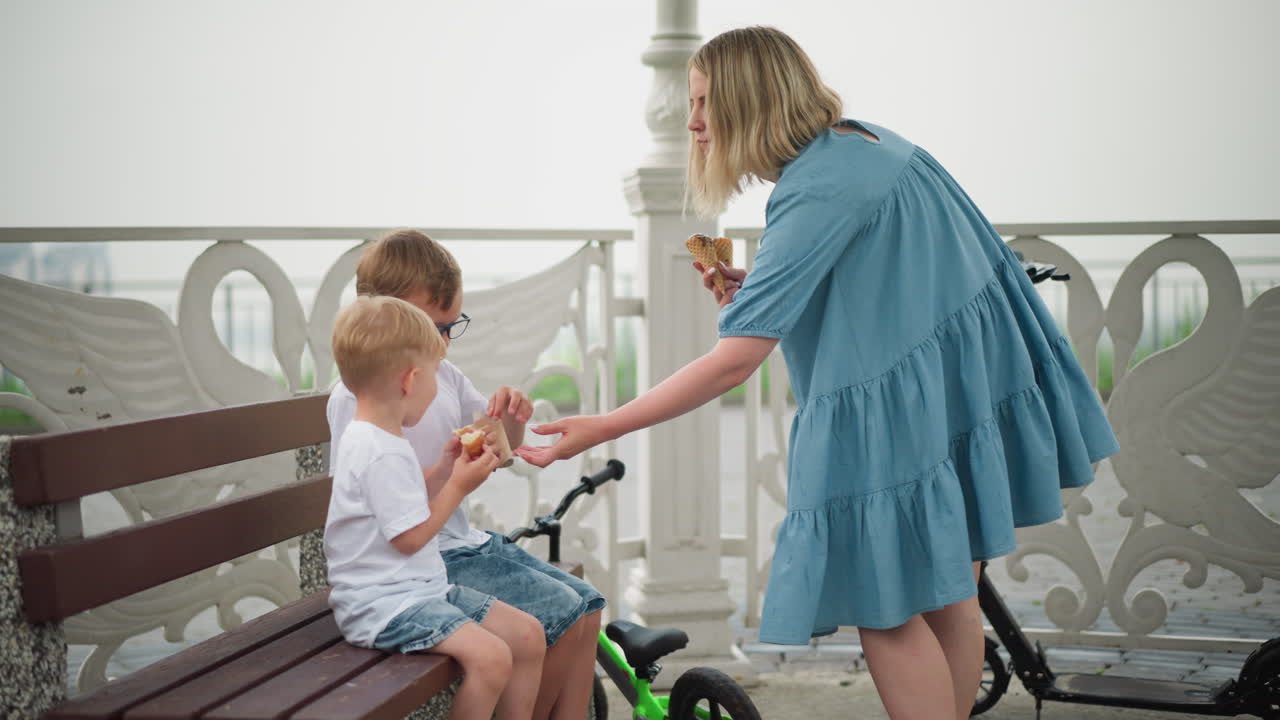 엄마는 오른손에 아이스크림 코너를 들고 큰 아들에게  한 봉투를 건네고, 어린 아이는 열렬하게 아이스크림을 잡으려고 손을 고 있다.