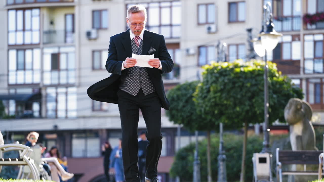 Full length portrait of a senior businessman who finds good news on paper walking in the city. Middle aged person in black suit becomes satisfied after reading outdoors. Slow motion.