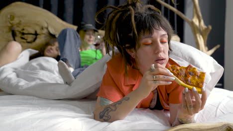 young woman with piercings and tattoos eating pizza while her friends are behind drinking wine and relaxing