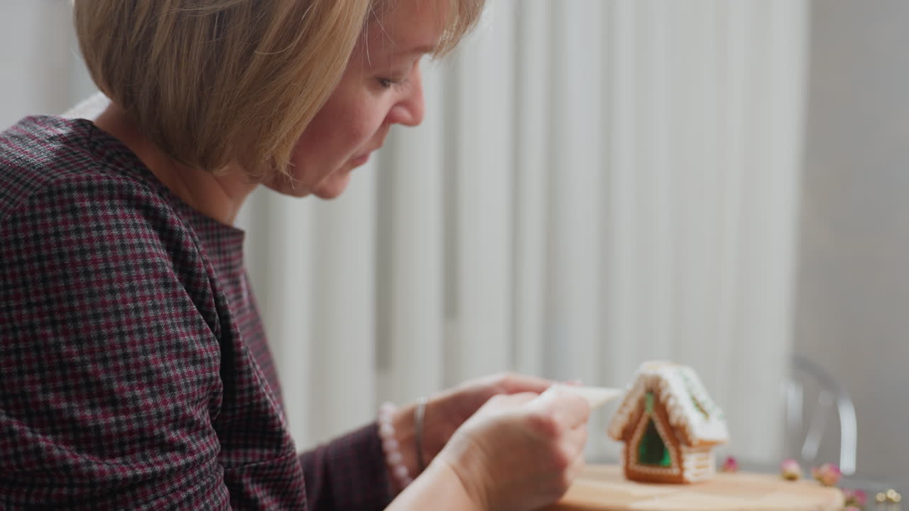 신선하게 구워진 진저브레드 집을 얼음으로 준비하는 여성의 측면 뷰, 그녀는 섬세한 세부 사항을 장식하기 전에 정밀성을 보장하여 아이싱 봉지를 조정합니다.