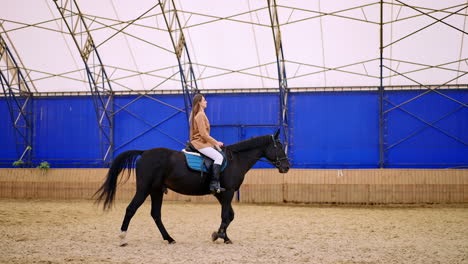 horse riding inside the drill-hall. long-haired woman in beige jacket and white pants rides a beautiful calm black horse.