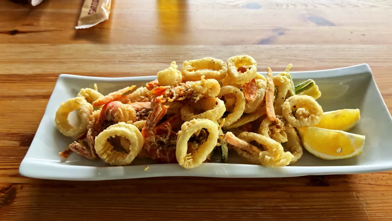 Riprese ravvicinate di calamari fritti serviti su un piatto da ristorante con spicchi di limone. Anelli dorati, panatura croccante e impiattamento pulito evidenziano la presentazione del piatto di mare e l'atmosfera culinaria