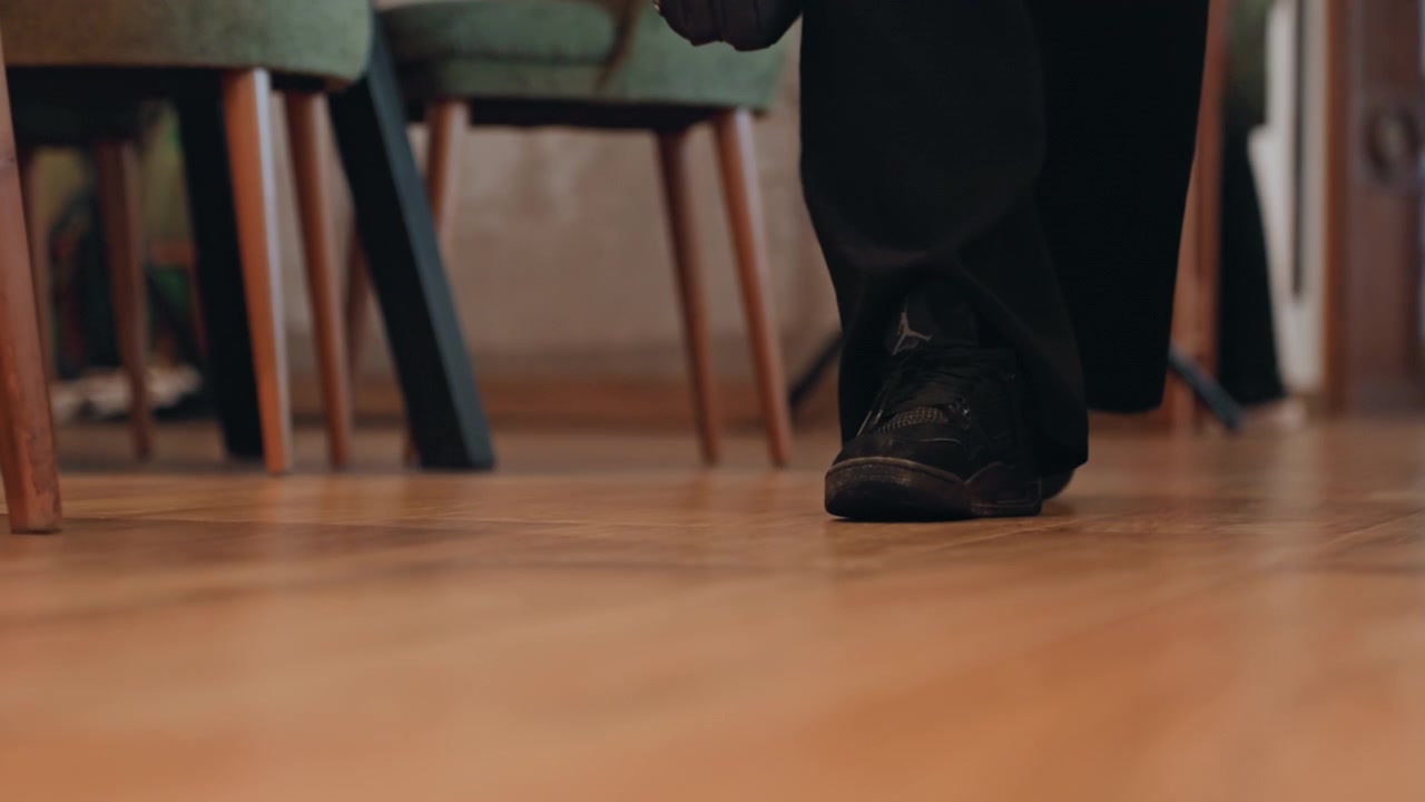 Closeup feet walking on wooden floor, photographer entering studio with tripod in background, black sneakers and wide trousers, slow rhythmic steps, polished parquet reflecting warm ambient light
