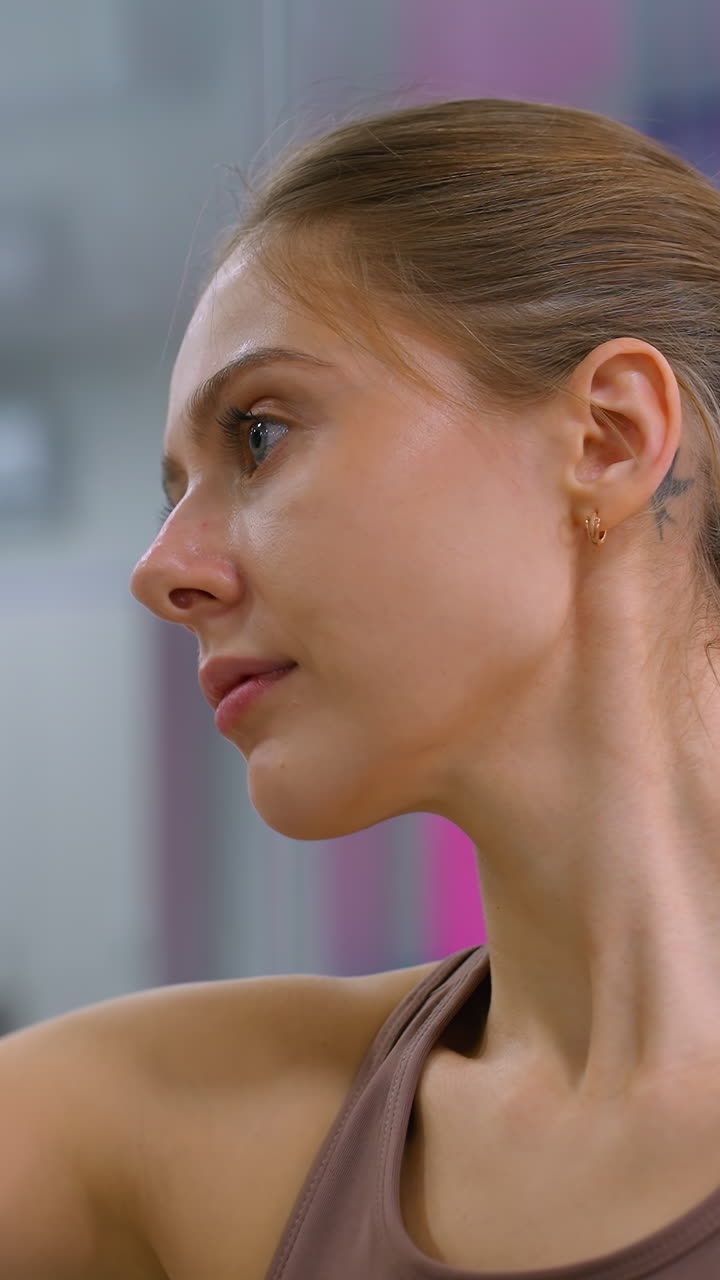 joven en forma pulveriza solución de limpieza en el vidrio y limpia el espejo con un paño azul, el espejo refleja el interior del gimnasio con equipos y cubiertas de ventanas rosas, expresión enfocada mientras asegura la limpieza