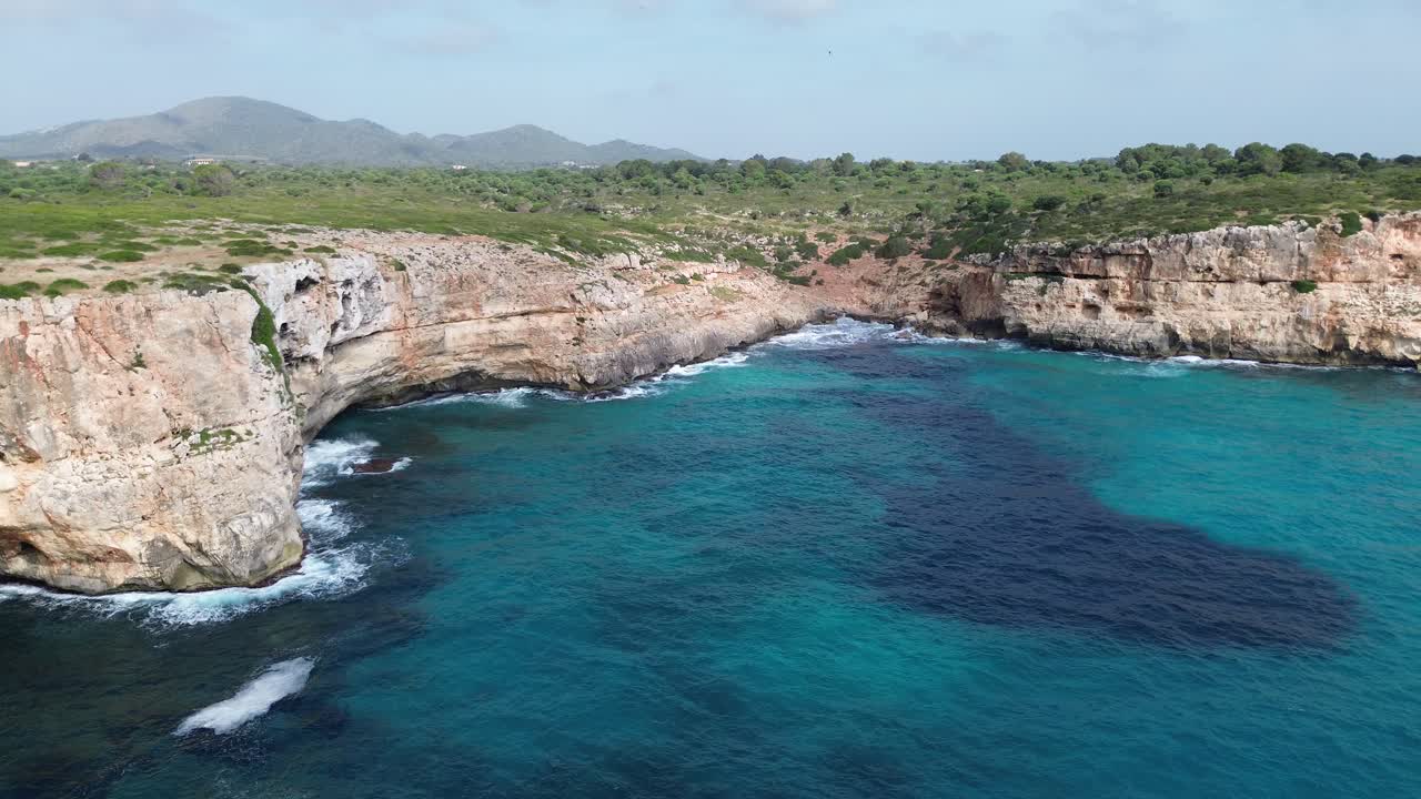 Imagens suaves de drone em 4K sobrevoando a costa de Maiorca em um dia ensolarado. Apresenta águas cristalinas, falésias rochosas, vegetação verde e montanhas ao longe para uma paisagem mediterrânea deslumbrante