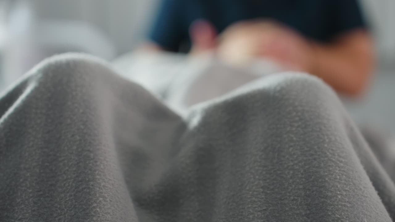 Woman covered in striped grey blanket lying on massage bed in bright spa room with faint blurred image of therapist massaging her during relaxing wellness session in serene tranquil environment