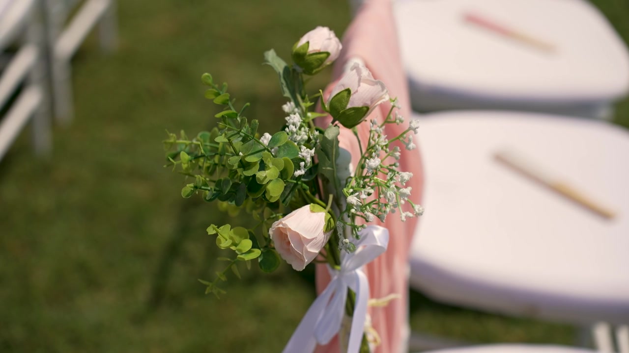 Gæster ankommer til et bryllupssted, hvor blomster er placeret på stole. Stolene er opstillet i en pæn række udenfor, omgivet af græs.