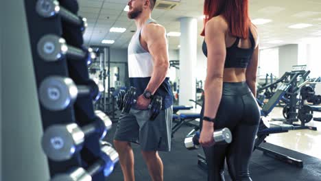 Close-up metal dumbbells with different weights in a gym. Slender female in black top and leggings is taking two dumbbells.