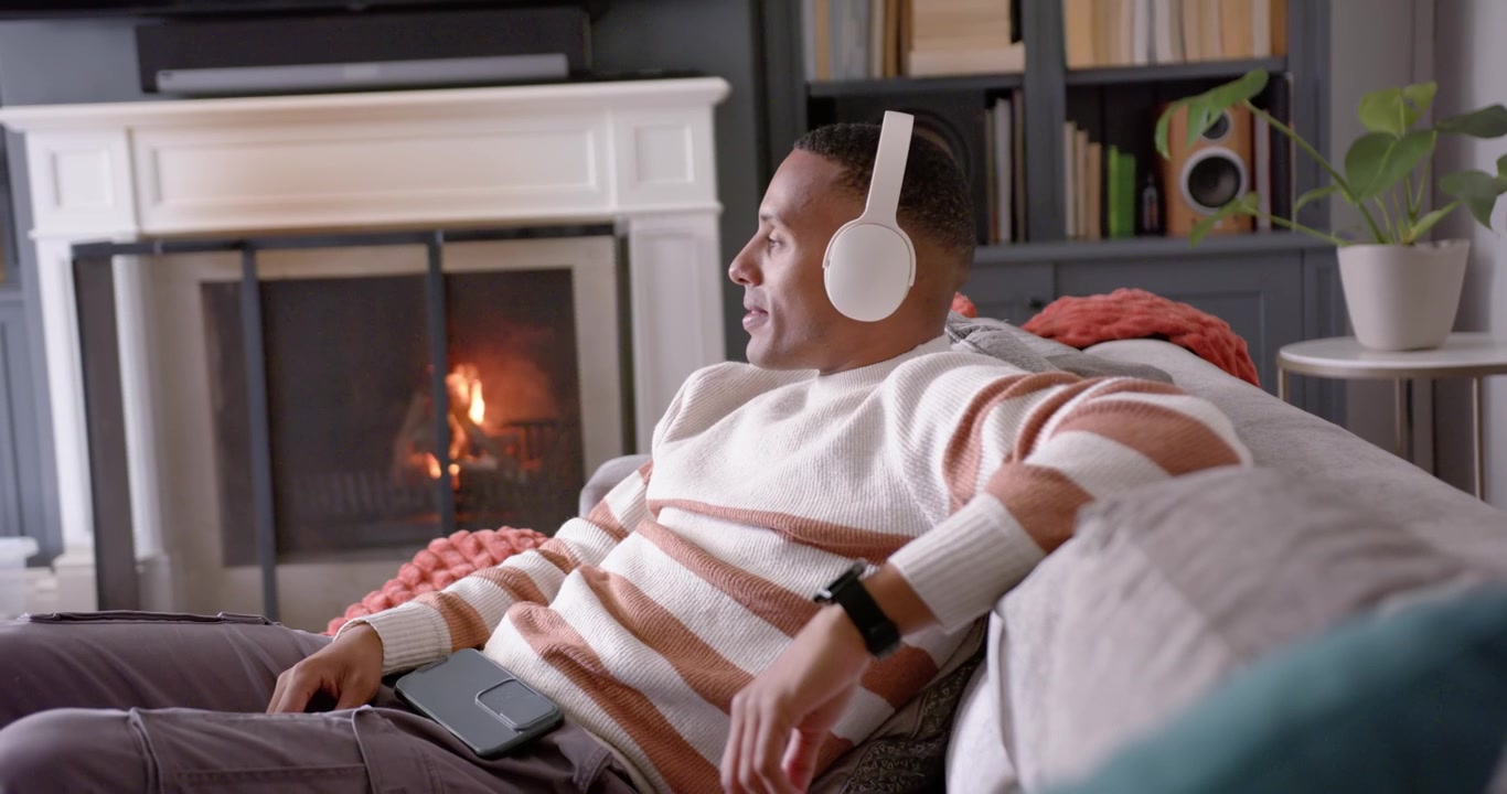 Uomo afroamericano adulto seduto sul divano a casa, con le cuffie ad ascoltare musica, mentre si rilassa con un dispositivo.