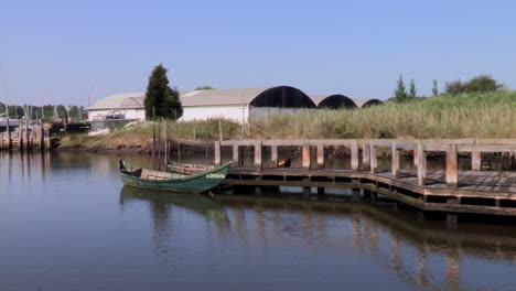 Fishing-pier-with-two-boats-and-wooden-walkway-in-the-middle-of-the-river,-warehouses-in-the-background-on-the-other-side-of-the-bank