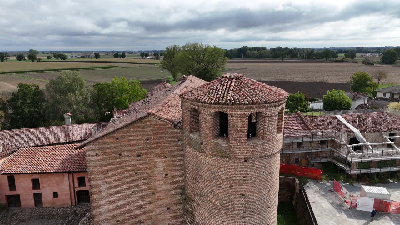 Castello di Calendasco a Calendasco, provincia di Piacenza, Italia, che mostra la torre medievale in mattoni, i tetti in terracotta, i ponteggi per il restauro e i terreni agricoli pianeggianti sotto nuvole drammatiche, ripresa con drone in orbita