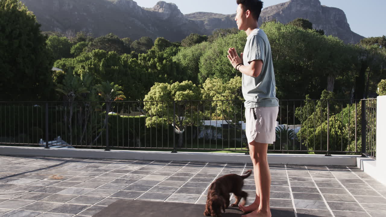 Asian man practicing yoga on patio with playful puppy, enjoying nature