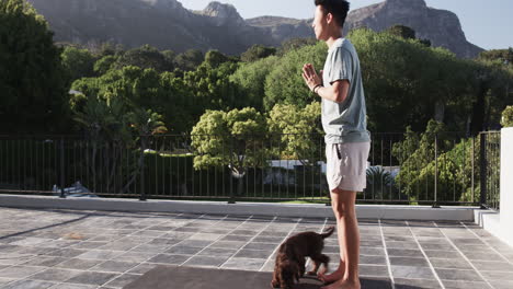 asian man practicing yoga on patio with playful puppy, enjoying nature
