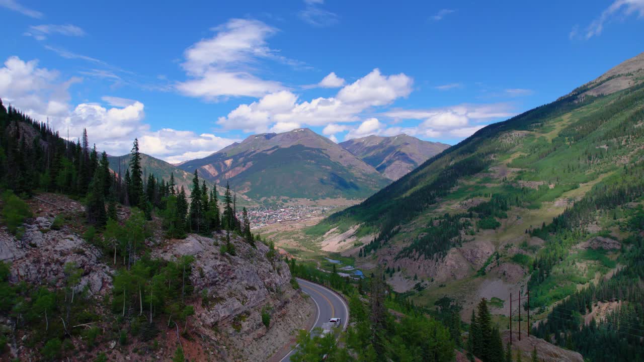 Ripresa aerea della storica città mineraria di Silverton, in Colorado, in una luminosa giornata estiva, con foreste lussureggianti e cime imponenti tutt'intorno. Delle auto percorrono la ripida strada tortuosa che conduce alla città di montagna.