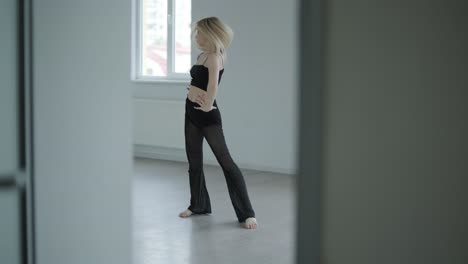 fit girl in black clothes performing contemporary. female dancer shows flowing motions, body and hands waves at white room. modern ballet dance choreography. young woman is moving smoothly