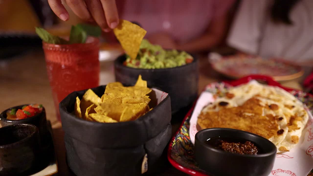A person grabs nacho chips and dips them into guacamole at a restaurant table with Mexican food, fresh salsa, and a red cocktail. Warm, inviting lighting and a shallow depth of field create a cozy dining atmosphere