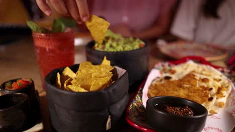 a person grabs nacho chips and dips them into guacamole at a restaurant table with mexican food, fresh salsa, and a red cocktail. warm, inviting lighting and a shallow depth of field create a cozy dining atmosphere