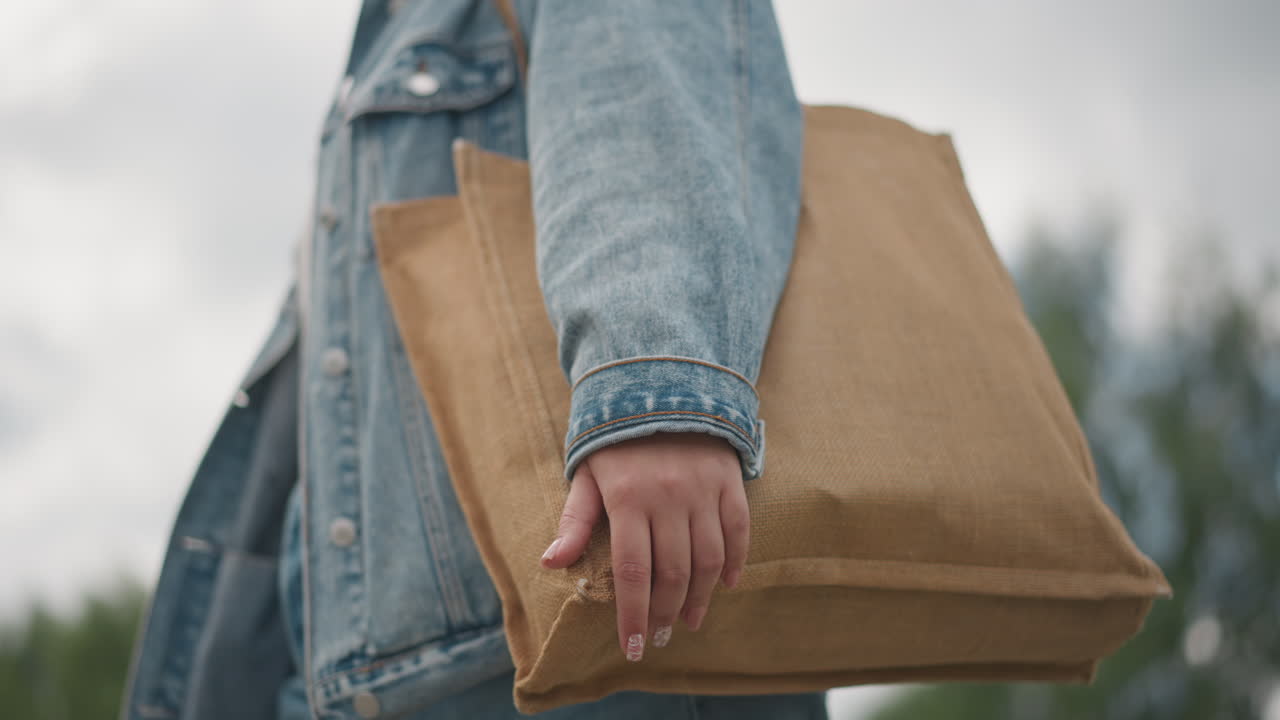Frau in Jeansjacke hält eine volle Einkaufstasche. Nahaufnahme einer Hand, die eine Jutetasche vor bewölktem Himmel festhält. Fester Schritt durch den Park. Ein strukturierter Sack, prall gefüllt mit Lebensmitteln. Praktischer Alltagsbegleiter und nachhaltig.