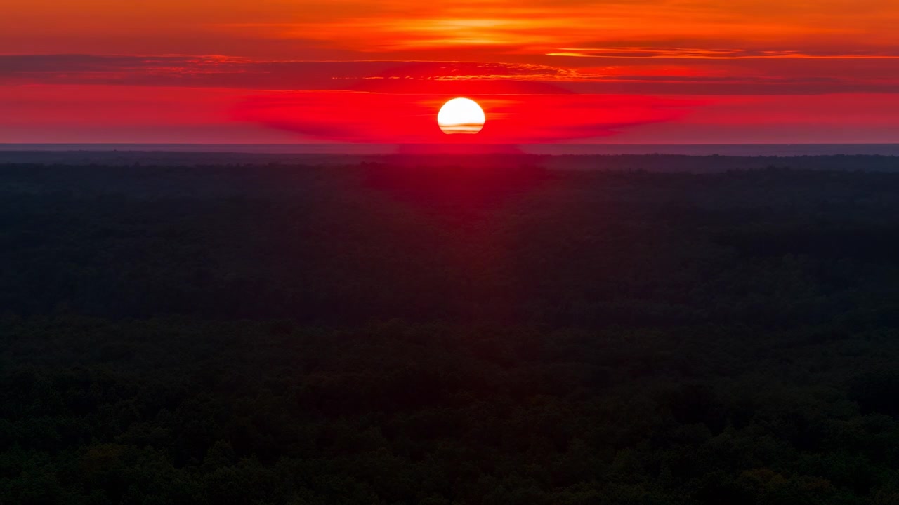 Den klare sol synker fra åben himmel til horisonten over et udstrakt skovkronetag, mens lagdelte røde, orange og lyserøde skyer intensiveres over det rolige aftenlandskab.
