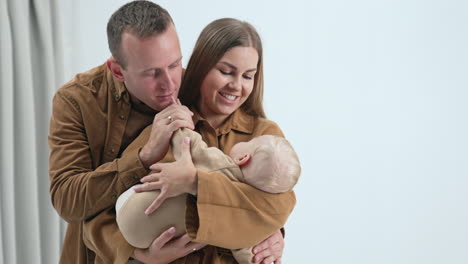 smiling long-haired woman holding a child in hands. father standing behind her kisses kid's hand tenderly. white backdrop.