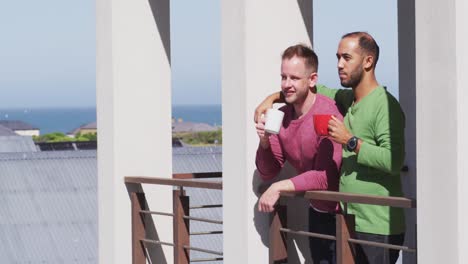 pareja de hombres homosexuales multiétnicos bebiendo café en el balcón al sol