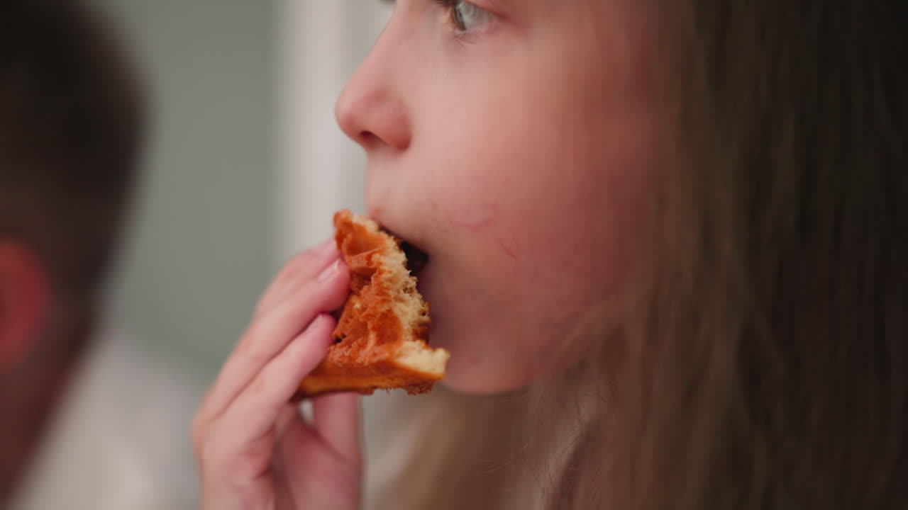 Close up of cute child licking chocolate filling from cookie with joyful expression while sitting beside another child in background slightly blurred eating something
