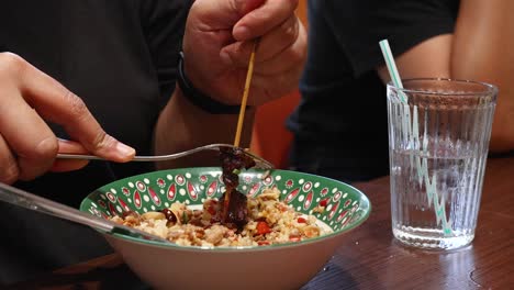 a person uses a fork and spoon to eat grilled beef skewer with rice in a patterned bowl, under warm indoor lighting, with a glass of water nearby