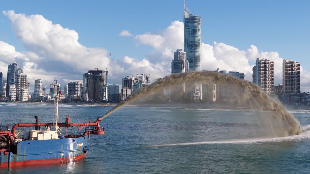 A dredging vessel pumps sand into the ocean against a city skyline under clear skies, highlighting coastal management