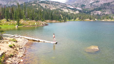 aerial drone flies over woman standing on the end of a fallen tree log in a clear blue water lake surrounded by mountains and thick pine tree forest in nederland colorado rocky mountains