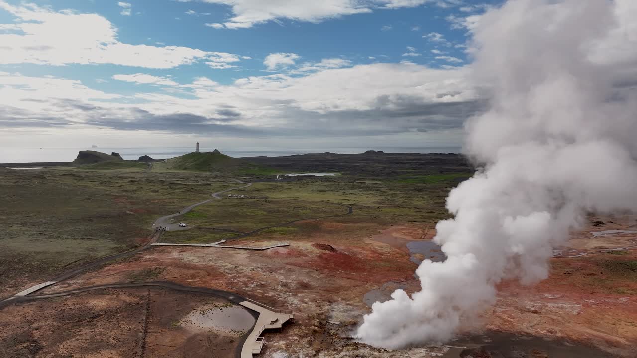Sobrevuelo aéreo de la zona geotérmica de Gunnuhver en Islandia con una potente fumarola de vapor emergiendo del paisaje volcánico, suelo mineral colorido, terreno accidentado y un cielo nublado dramático. Plano general
