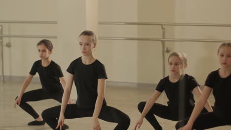 a group of young ballet students in black dancewear practicing positions in a spacious ballet studio with wooden flooring and wall-mounted barres. focused expressions and synchronized movements.