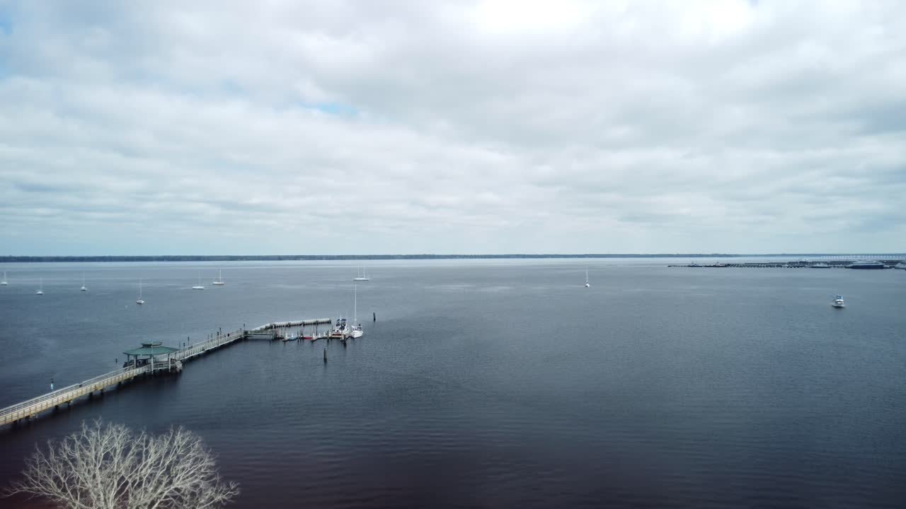 A moody drone shot flies over the river, soaring out over a pier on an overcast day, capturing the calm, misty atmosphere.